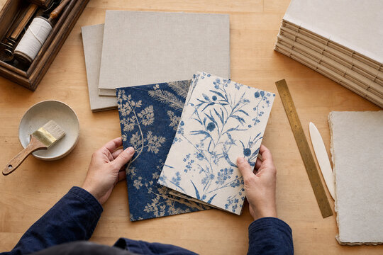 Overhead view of hands holding handmade papers with blue botanical prints on a wooden craft table with bookbinding tools and paper stacks.