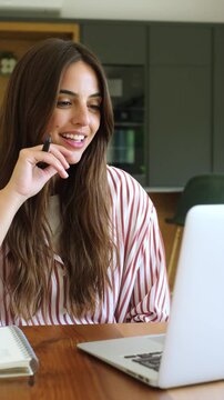 Smiling professional woman sitting at her desk, engaging in an online meeting, and speaking during a video conference while working from her modern home office kitchen with a laptop