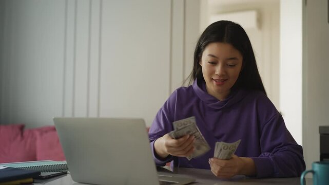 Asian woman counting cash beside a laptop, pleased with business profits