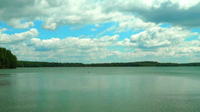 A large lake is the main focus as the sun shines down on the water. The surface is smooth and calm, showing the shape of clouds above. Trees line the shore, creating a natural border around the lake. 