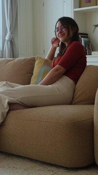 Peaceful young woman with glasses sitting on a comfortable couch in her living room, smiling and relaxing. Serene atmosphere of a quiet afternoon at home, showcasing a calm and content lifestyle