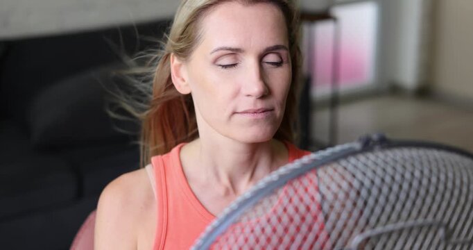 Woman enjoying wind from electric fan with eyes closed. Hair is blowing. Air conditioning and cooling at home on summer heat