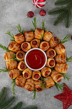 Christmas wreath of pigs in blankets with tomato sauce, garnished with fresh rosemary and sesame. Christmas appetizer on a stone background. Top view, copy space.﻿