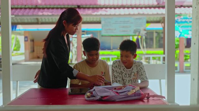 Smart Asian primary students studying and reviewing lessons with a helpful female teacher outside the classroom. Cheerful young boys learning together with school textbooks.