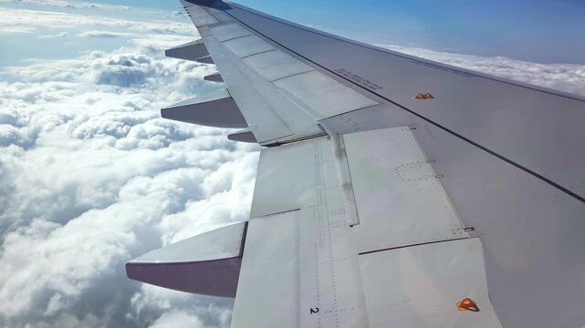 Airplane wing viewed from window flying above fluffy clouds in bright blue sky, showcasing the wing's flaps and ailerons in a dynamic aerial perspective