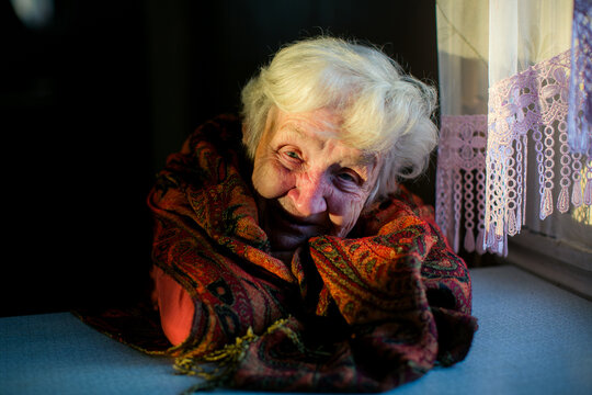 Portrait of a smiling elderly woman wrapped in a colorful woven shawl, sitting by a sunlit window with lace curtains in a cozy home interior.