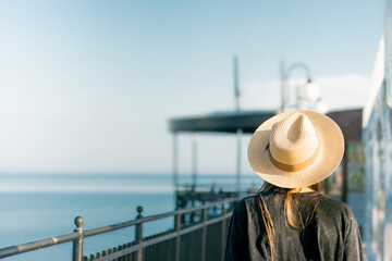 Woman wearing a wide-brimmed straw hat stands by a railing overlooking a calm sea with a clear sky and distant structures visible in the background © Anastasiia