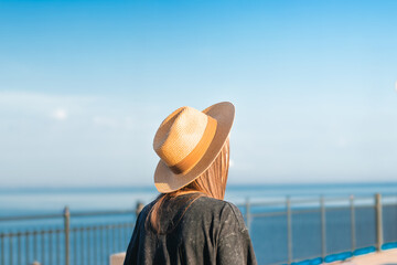 Woman with long brown hair wearing a straw hat stands by a railing overlooking a calm sea under a clear blue sky on a sunny day © Anastasiia