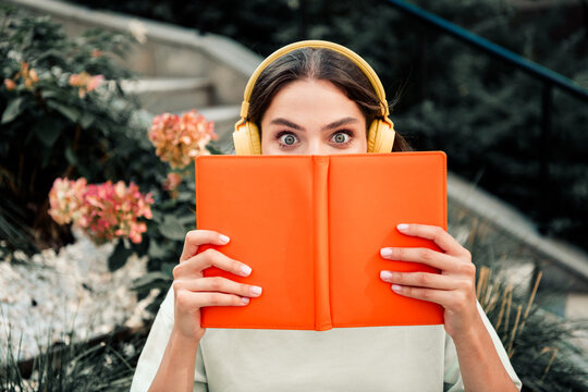 Young woman in yellow headphones peeks over an orange book with wide surprised eyes while reading outdoors in a garden setting