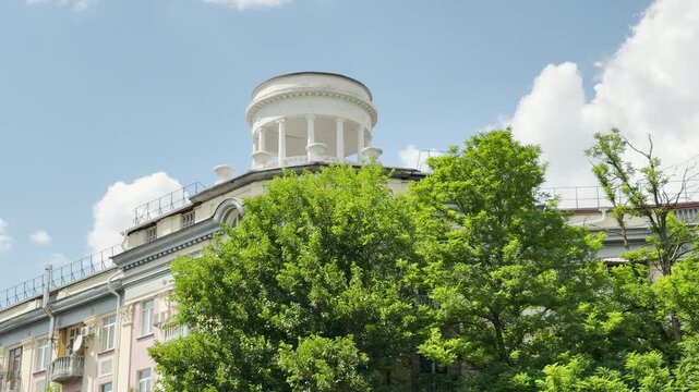 A beautiful classical building stands peacefully behind lush green trees under a bright blue sky in Simferopol, Crimea. This serene view evokes a feeling of calm nostalgia.