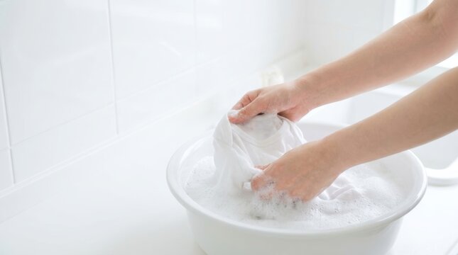 Hands washing a white garment in a basin filled with soapy water with tiled wall