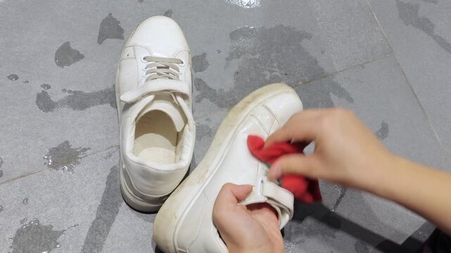 A dirty white sneaker is cleaned with a red sponge on gray tile flooring.