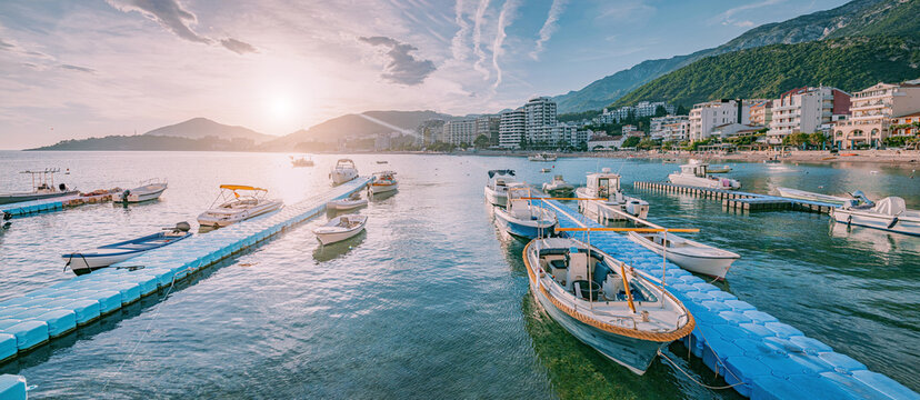 Boats moored at blue floating docks in Budva, Montenegro, with a coastal town, green mountains, and the Adriatic Sea at sunset