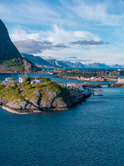 Fototapeta premium Tiny rocky islet in the Lofoten Islands, Norway, with turf roof cabins and rorbuer huts by a small pier, deep fjord and jagged mountains, summer daylight, scattered clouds.