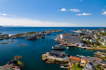 Obraz premium Aerial view of a Lofoten Islands town in Norway shows a low bridge, islets, rorbuer houses, fisheries, silos, and boats in a sheltered harbor under a clear summer sky.