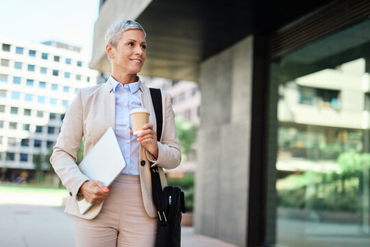 Portrait of a young businesswoman woman walking down the street, holding a laptop computer and a coffee cup, surrounded by moder corporate office buildings