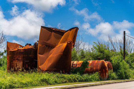 View of rusted and damaged metal tanks standing in an overgrown field under a bright blue sky in San Jose, Tinian, Northern Mariana Islands.