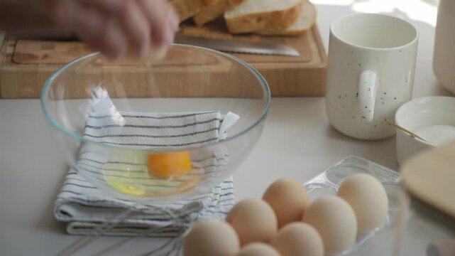 Hand cracking eggs into glass bowl on kitchen counter, with bread, mug and eggs nearby. Concept of breakfast preparation and home cooking