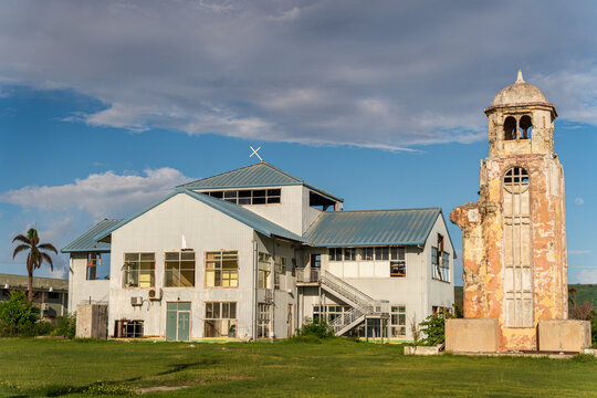 View of the historic San Jose Catholic Church with its weathered bell tower and adjacent modern building under a blue sky in San Jose, Tinian, Northern Mariana Islands.