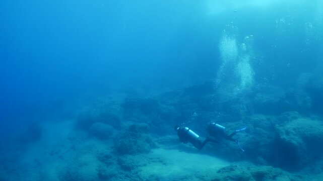  scuba divers exploring around a reef underwater deep blue water big rocks and bubbles ocean scenery 
