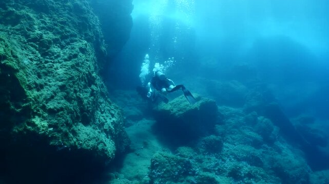  scuba divers exploring around a reef underwater deep blue water big rocks and bubbles ocean scenery 