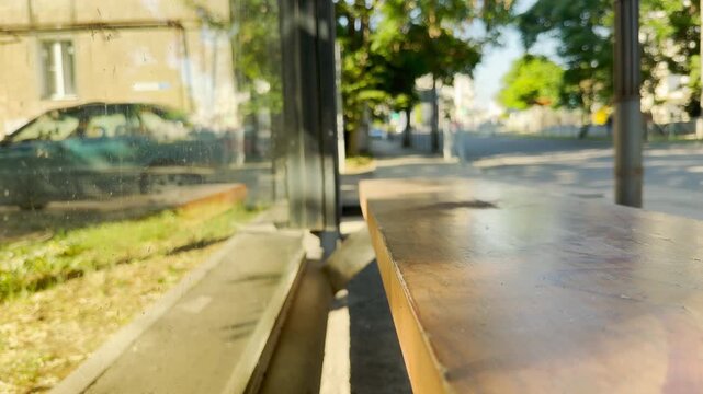 An empty wooden bench waits inside a glass shelter at a sunlit bus stop in Simferopol, Crimea. This quiet scene brings a lonely, peaceful, and nostalgic feeling.