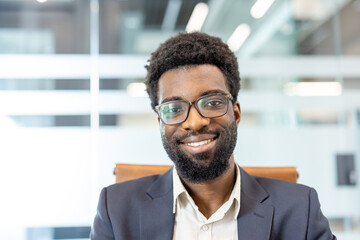 Fototapeta premium Young adult professional male wearing eyeglasses and a suit jacket. Smiling confidently while looking at the camera. Portraying success and a friendly business demeanor in a modern office environment