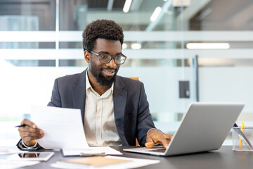 Obraz premium Confident young entrepreneur using mobile computer and holding paper documents, focusing on business tasks in a modern workplace setting, working with technology and information