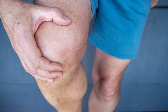 A man holding his right knee with his right hand, wearing short blue shorts, representing joint or muscle pain, injury, illness and medical symptoms.