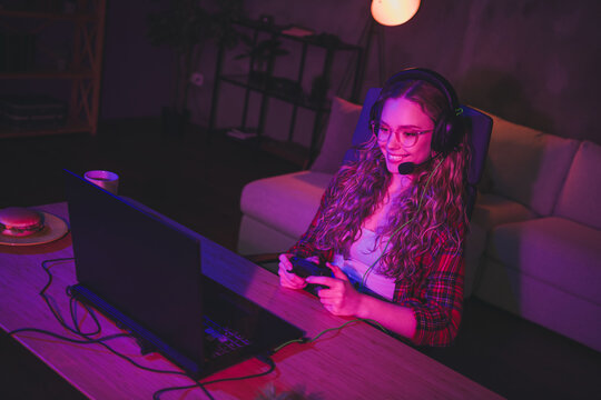 Photo of gamer blonde teen lady hold joystick play netbook strategy game wear plaid shirt late night room indoors pink blue neon light