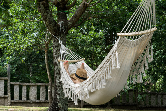 Side view of a woman completely relaxed, wearing a straw hat and lying in a comfortable white fringed hammock hung in a green garden.