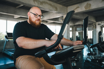 Obraz premium Man exercise bike gym fitness workout indoor bearded eyeglass overweight man using stationary exercise bike in modern gym for cardio training