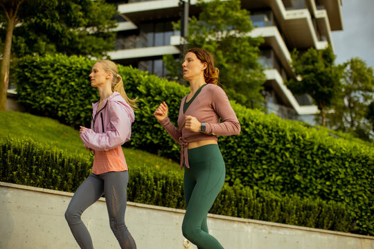 Two women jog along a path in a city with modern buildings in the background