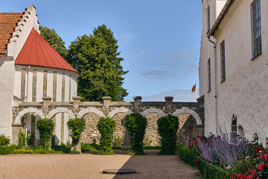 View of the historic Bosjokloster Abbey complex featuring a stone arched walkway, red-roofed building, and lush garden flowers in Bosjokloster, Skane County, Sweden.