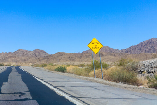 Yellow diamond-shaped warning sign reading watch for rocks stands next to a desert highway with mountains behind near Yuma, Arizona; copy space