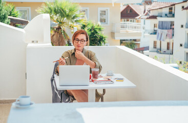 Naklejka premium Joyful female painter sitting on balcony creating artwork during seaside vacation. Relaxed woman smiling, enjoying creative process and sunny coastal atmosphere, combining leisure, art and inspiration