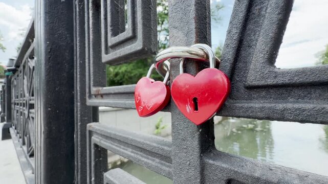 Two red heart padlocks are locked tightly onto a metal bridge railing over a river. They represent a couple's eternal love, romantic hope, and joyful commitment.