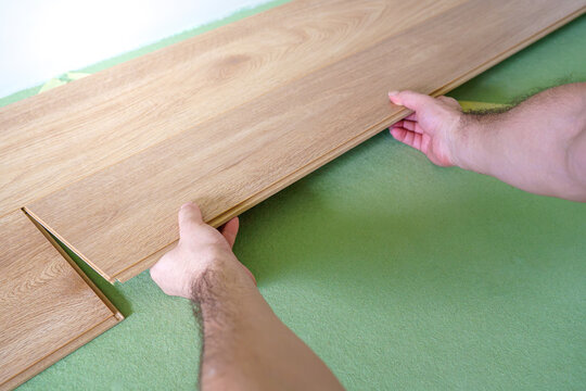 Close-up of a craftsman's hands joining oak laminate wood planks together over a green underlay foam layer during home renovation.