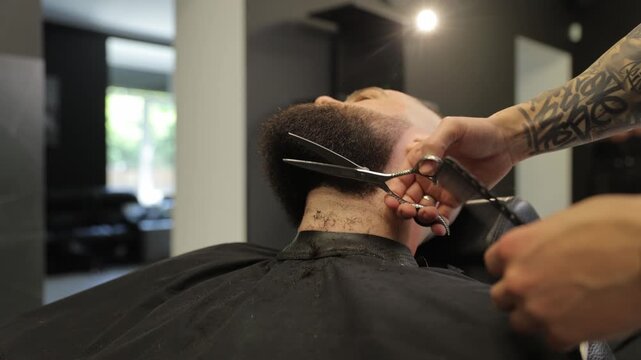 Tattooed barber trimming man's beard with scissors