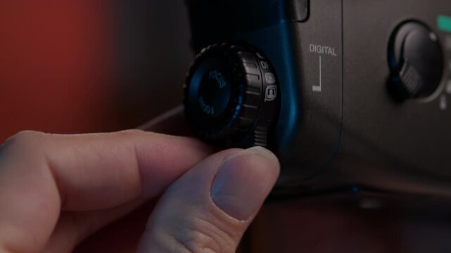 Professional cinematographer hand manipulating focus mode dial on digital video camera, switching between settings and preparing equipment for shoot in dimly lit studio, close-up, slow motion