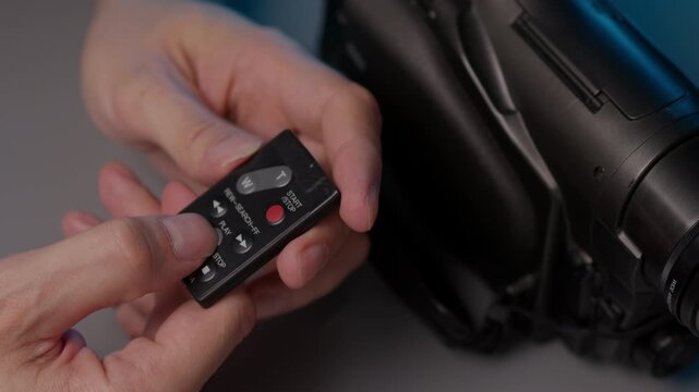 Human hands holding and operating small black remote control for retro camcorder, pressing buttons to navigate, play, search through vintage video recordings from past, close-up, slow motion
