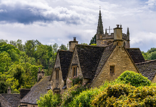 Charming view of St Andrew Church spire rising above classic Cotswold stone cottages and lush greenery in Castle Combe, England.