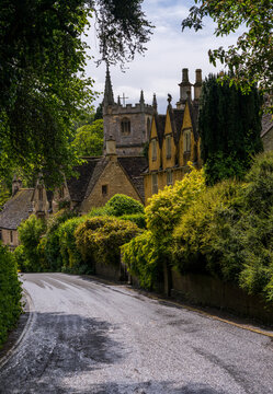 Charming view of St Andrew Church spire rising above classic Cotswold stone cottages and lush greenery in Castle Combe, England.