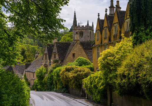Charming view of St Andrew Church spire rising above classic Cotswold stone cottages and lush greenery in Castle Combe, England.