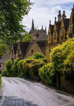 Charming view of St Andrew Church spire rising above classic Cotswold stone cottages and lush greenery in Castle Combe, England.