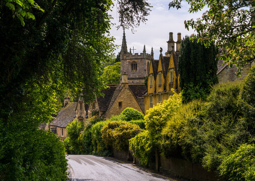 Charming view of St Andrew Church spire rising above classic Cotswold stone cottages and lush greenery in Castle Combe, England.