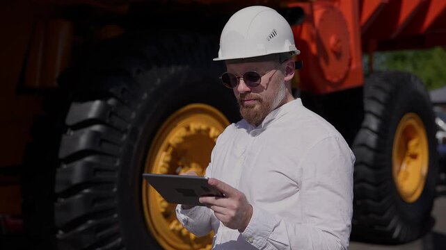 Worker construction engineer using tablet to inspect heavy machinery at outdoor worksite during daytime.