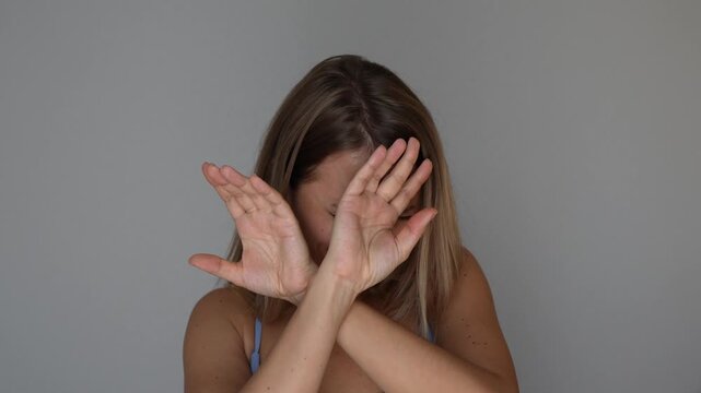 Young blonde woman rejects something unpleasant and frightening, spreading her hands in denial and trying to hide behind them in a fear and avoidance concept on a grey background