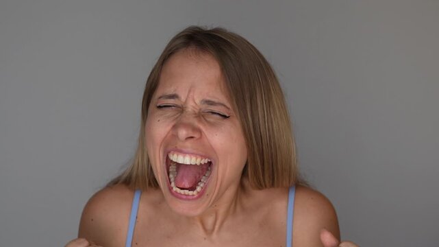Young blonde woman screams loudly to release emotions, expressing intense frustration, anger or emotional overflow in a powerful emotional portrait on a grey background 