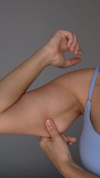 Young blonde woman touches the soft lower part of her upper arm, showing flabby underarm muscle in a body-related close-up concept on a grey background